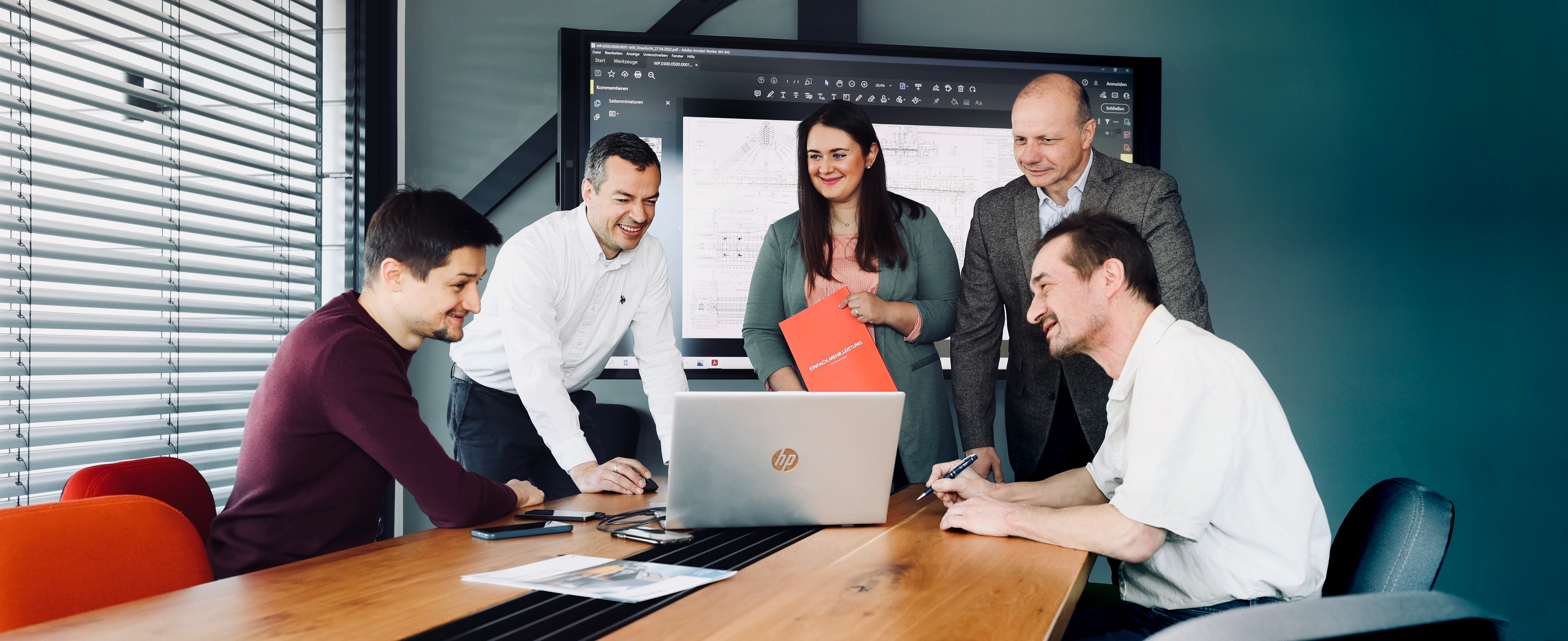 Team aus fünf Personen im Besprechungsraum, gemeinsam am Laptop mit Bildschirm im Hintergrund