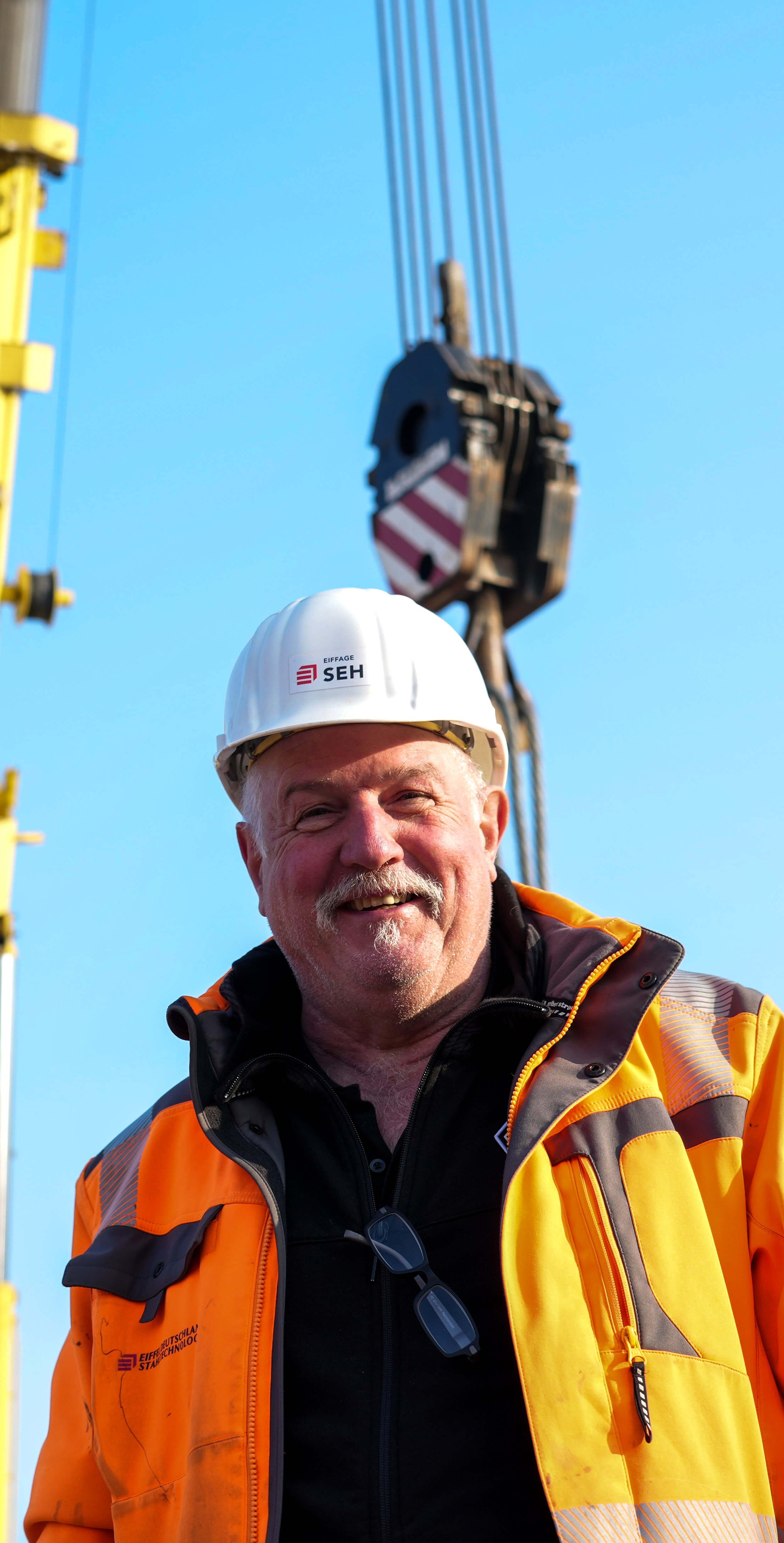 Älterer Mitarbeiter in orangener Schutzkleidung mit Helm, lacht in die Kamera – im Hintergrund ist ein Teil eines Krans erkennbar