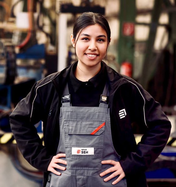 Junge Mitarbeiterin vor industriellem Hintergrund in Arbeitskleidung mit Latzhose, lächelt freundlich in die Kamera 