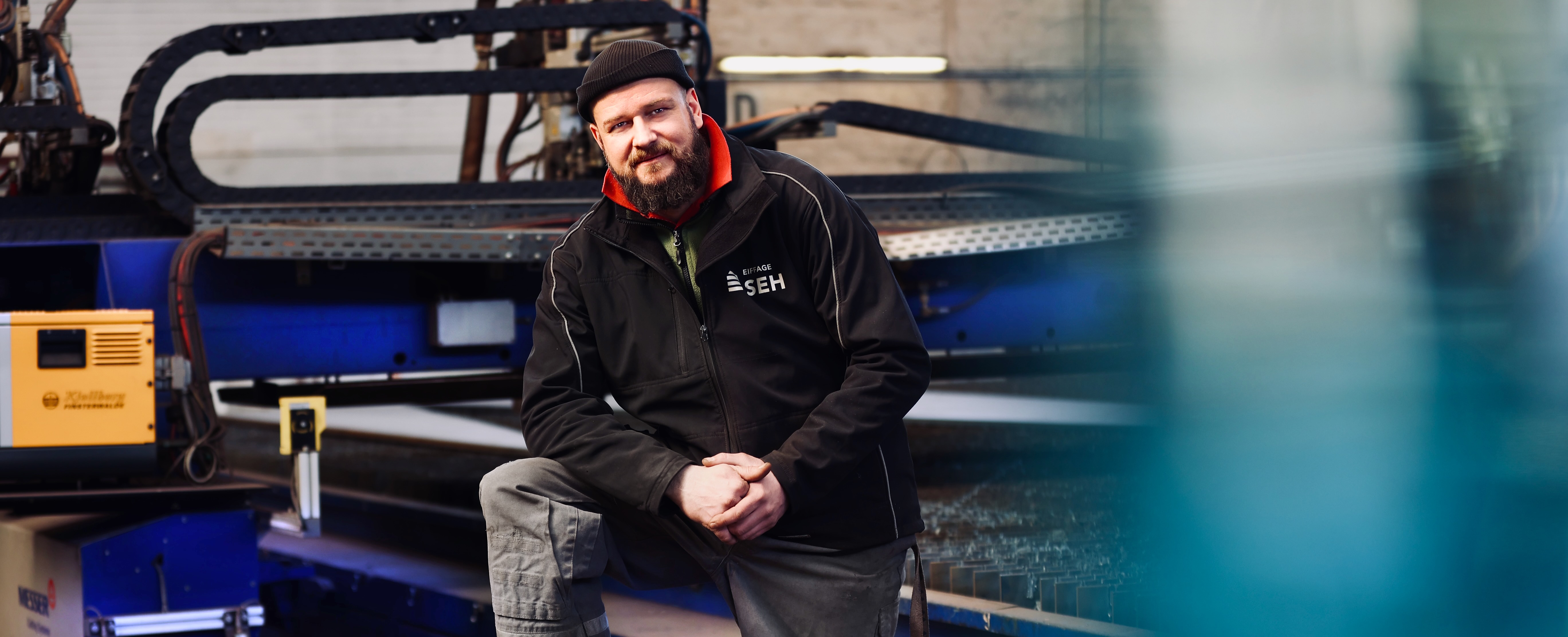 Mitarbeiter mit Bart in legerer Arbeitskleidung, sitzt auf einer blauen Maschine oder einem Fahrzeug